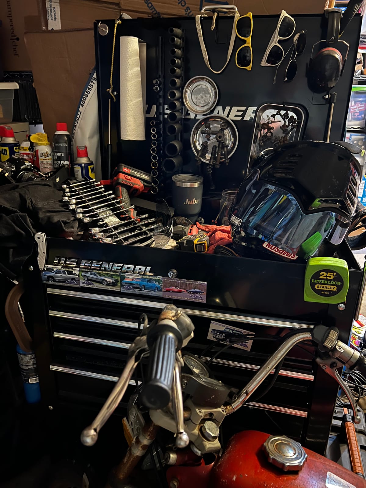 A large multi-drawer toolbox with various tools scattered about on top along with a motorcycle helmet and a red moped nearby.