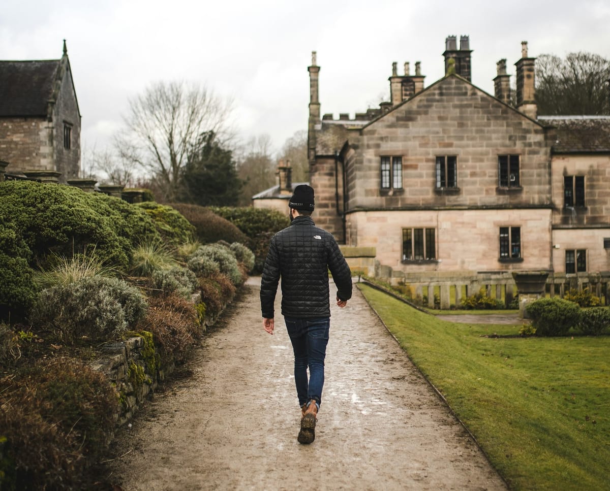 A man in dark pant, dark jacket, and a dark beanie walks down a muddy path between stone-walled houses.